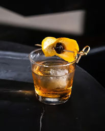 Classic whiskey Old Fashioned in a short rocks glass with a large ice cube, glowing amber liquid and an orange peel twist with a cherry on a bamboo pick, perched on a dark marble bar top.