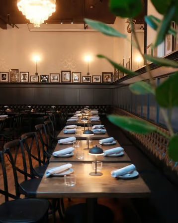 Cozy upscale bistro interior with a long row of wooden tables set with rolled napkins and small table lamps along a tufted banquette, warm wall sconces and framed photos, chandelier and leafy plant in the foreground.