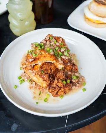 Crispy fried chicken biscuit topped with creamy sausage gravy and chopped green onions on a white plate — brunch comfort food on a cafe table