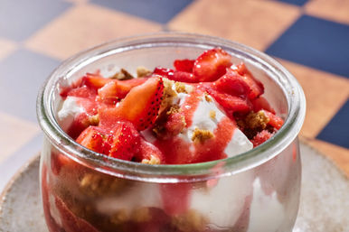 Bright strawberry parfait in a small glass jar—layers of creamy yogurt, fresh sliced strawberries, strawberry sauce and crunchy granola crumbs on a ceramic plate over a blue-and-tan checkered table.