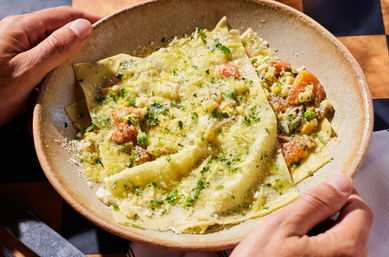 Sunlit hands holding a rustic bowl of fresh wide pasta sheets layered over shrimp and vegetable filling, drizzled with olive oil and sprinkled with grated Parmesan and chopped parsley.