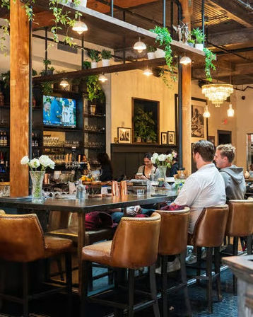 Modern industrial-chic gastropub interior with leather bar stools at a long communal table, hanging plants and pendant lights, patrons seated, TV on the back wall and vases of fresh flowers.