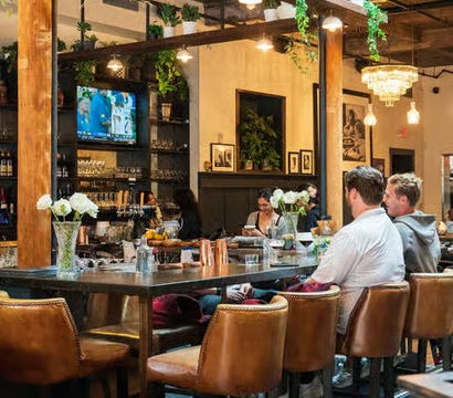 Modern industrial-chic gastropub interior with leather bar stools at a long communal table, hanging plants and pendant lights, patrons seated, TV on the back wall and vases of fresh flowers.