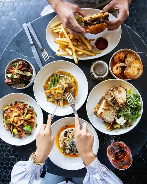 Overhead shot of a downtown bistro table with hands sharing a burger and fries, plated pastas, grilled sandwiches, salads and a glass of rosé