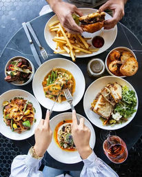 Overhead shot of a downtown bistro table with hands sharing a burger and fries, plated pastas, grilled sandwiches, salads and a glass of rosé