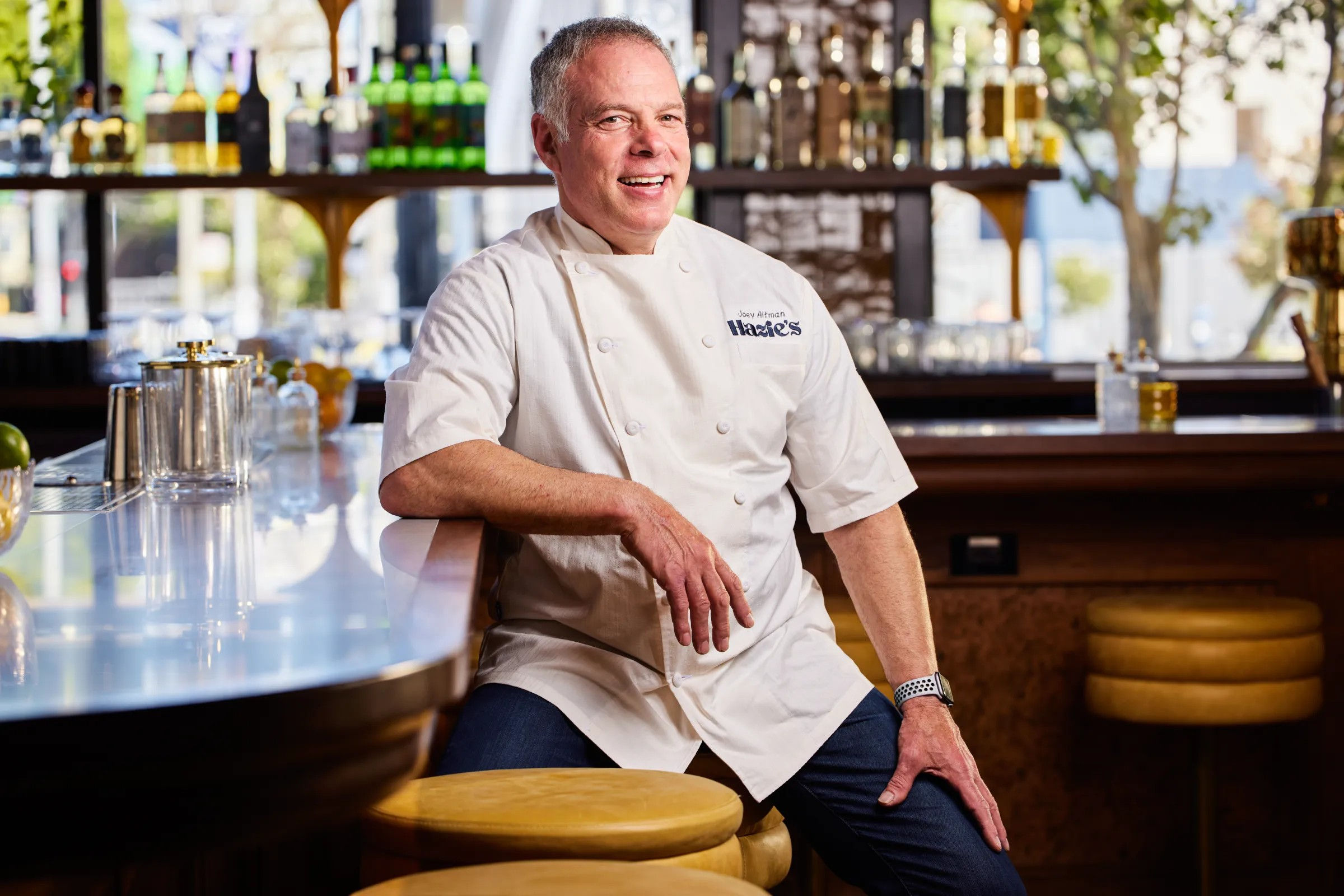 Smiling chef in white coat leaning on a shiny barstool at a bright urban restaurant bar with bottles on back shelves and warm natural light
