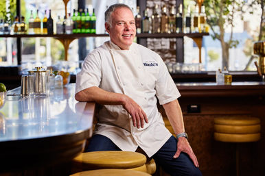 Smiling chef in white coat leaning on a shiny barstool at a bright urban restaurant bar with bottles on back shelves and warm natural light