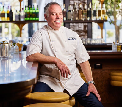 Smiling chef in white coat leaning on a shiny barstool at a bright urban restaurant bar with bottles on back shelves and warm natural light