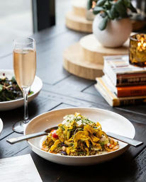 Bright ribbon pasta with roasted vegetables and herbs in a white bowl on a dark wood cafe table, accompanied by a glass of sparkling wine, a side salad, stacked books and a potted succulent in soft natural light.