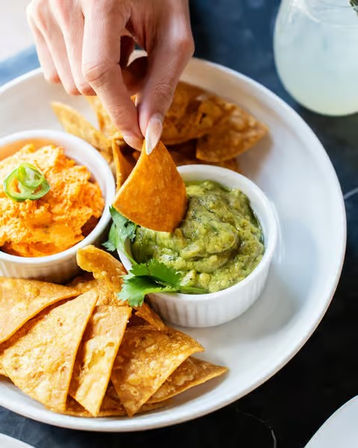 Hand dipping a tortilla chip into creamy guacamole on a white plate with extra chips and a spicy orange cheese dip topped with jalapeño — Mexican-style appetizer
