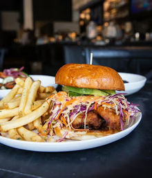 Crispy fried chicken sandwich on a shiny brioche bun topped with colorful cabbage-carrot slaw and avocado, served with seasoned French fries on a white plate in a casual restaurant