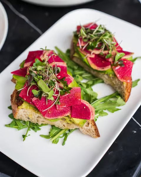 Open-faced avocado toast topped with bright watermelon radish slices and microgreens on a bed of arugula, served on a white rectangular plate.