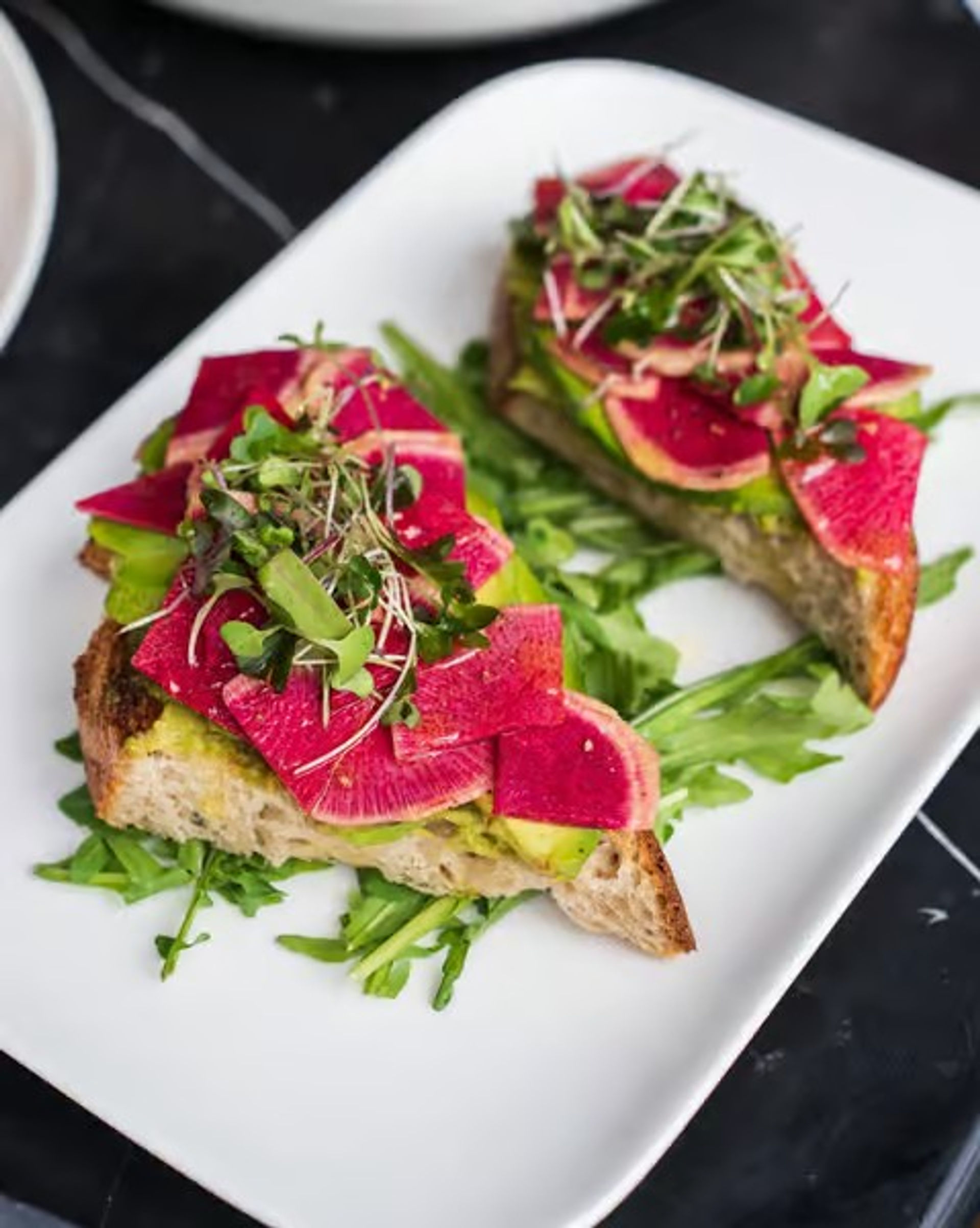 Open-faced avocado toast topped with bright watermelon radish slices and microgreens on a bed of arugula, served on a white rectangular plate.