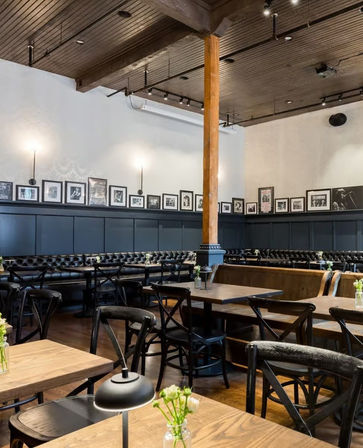 Cozy urban cafe interior with wooden tables, black cross-back chairs, tufted leather banquette seating, framed photo gallery wall, exposed wood column and dark wood ceiling.