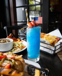 Tall bright blue frozen cocktail in a highball glass, garnished with strawberries, a striped straw and small American flag, on a casual restaurant table with fries, a sandwich and a side salad.