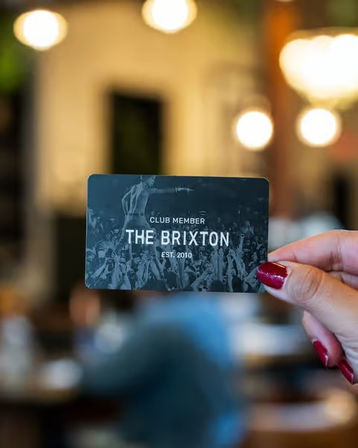 Hand with red nail polish holding a dark club membership card featuring a concert crowd image, blurred cozy cafe/bar interior with warm bokeh lights in the background