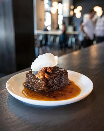 Square chocolate cake in caramel sauce topped with walnuts and a dollop of whipped cream on a white plate, sitting on a dark wood counter with a blurred cafe background