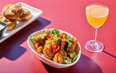 Bowl of crispy chili-glazed fried chicken tossed with fresh cilantro and sliced red chilies, paired with golden dinner rolls on a platter and a bright citrus cocktail in a stemmed glass on a red tabletop.