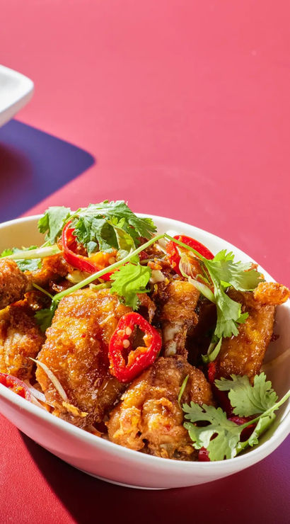 Bowl of crispy chili-glazed fried chicken tossed with fresh cilantro and sliced red chilies, paired with golden dinner rolls on a platter and a bright citrus cocktail in a stemmed glass on a red tabletop.