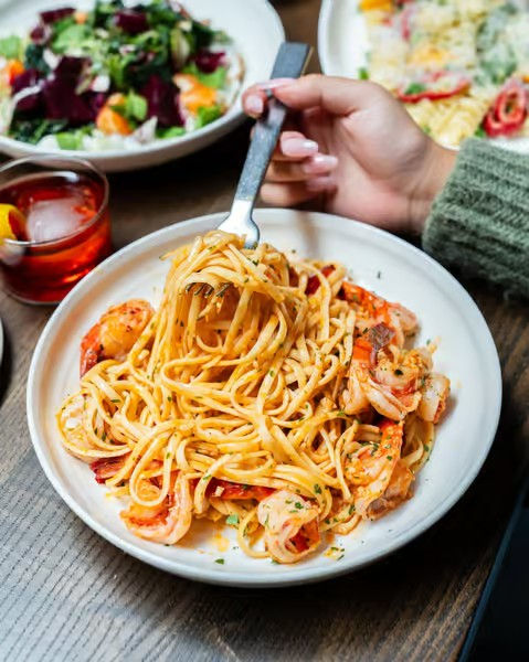 Shrimp linguine tossed in creamy tomato sauce on a white plate, fork twirling noodles at a cozy bistro table with salad and cocktail nearby.