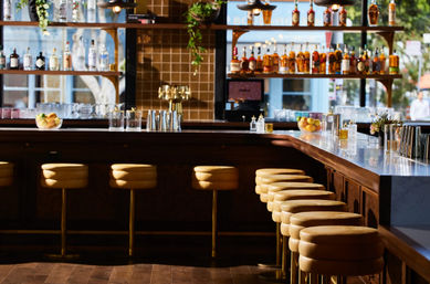 Sunlit modern cocktail bar interior with a row of tan leather stools along a marble counter, backlit liquor shelves, brass taps and citrus bowls, inviting urban vibe.