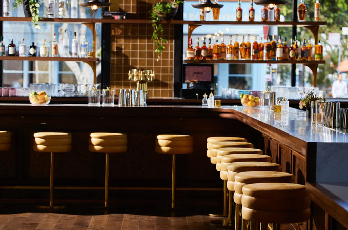 Sunlit modern cocktail bar interior with a row of tan leather stools along a marble counter, backlit liquor shelves, brass taps and citrus bowls, inviting urban vibe.