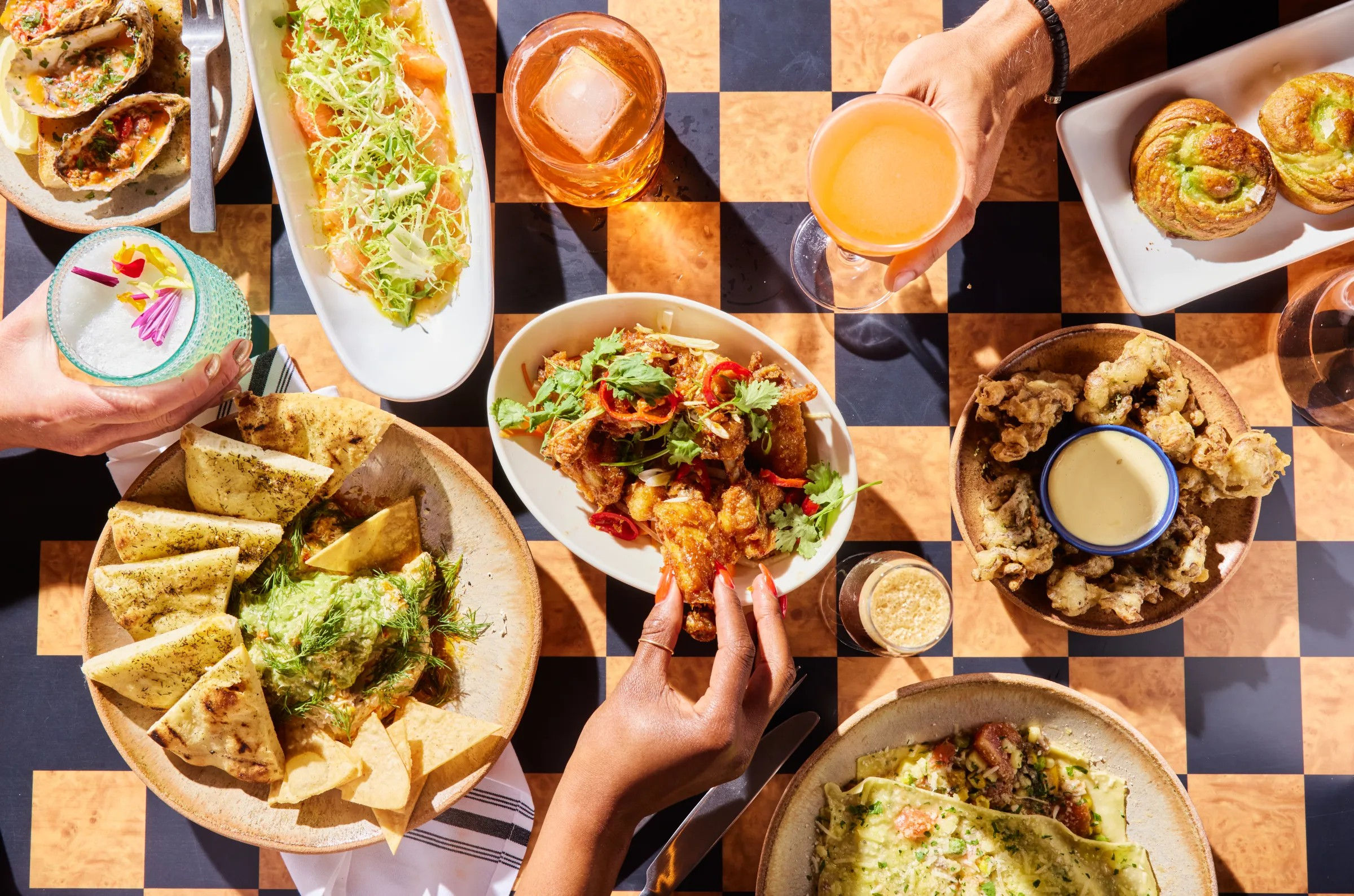 Sunlit overhead shot of a vibrant shared brunch on a checkered patio table: pita and herbed dip, fried chicken bites with cilantro and chilies, crispy fried mushrooms with dipping sauce, stuffed flatbread, savory rolls and colorful craft cocktails, hands reaching for bites.