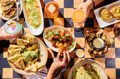 Sunlit overhead shot of a vibrant shared brunch on a checkered patio table: pita and herbed dip, fried chicken bites with cilantro and chilies, crispy fried mushrooms with dipping sauce, stuffed flatbread, savory rolls and colorful craft cocktails, hands reaching for bites.