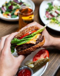 Close-up of hands holding a juicy cheeseburger with melted cheddar, lettuce and tomato on a sesame bun, served with pickle spears, ketchup and side salads on a wooden restaurant table.
