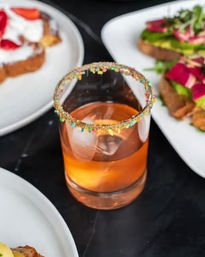 Sprinkle-rimmed cocktail in a lowball glass with a large ice cube and amber drink, surrounded by avocado toast and sweet toast on white plates — colorful brunch scene.