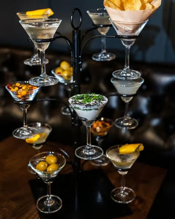 Martini glass tower at a stylish bar — tiered martini glasses on a black metal stand holding cocktails and garnishes like olives, lemon twists, chips and a creamy dip.