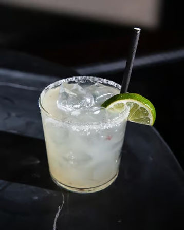 Chilled margarita-style cocktail in a short rocks glass with salted rim, ice cubes, black straw and lime wedge on a glossy bar top