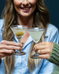 Two people clinking martini glasses with pale cocktails, one garnished with an olive, smiling person blurred in the background for a cozy bar/happy hour vibe.