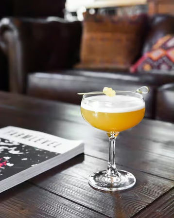 Citrus foam cocktail in a coupe glass on a rustic wooden coffee table, garnished with a small twist, with a leather sofa and magazine in a cozy lounge background.