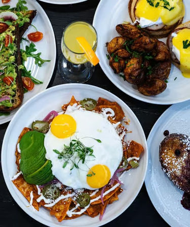 Overhead shot of a colorful brunch spread: chilaquiles topped with two sunny-side eggs, sliced avocado, jalapeños and crema, avocado toast with cherry tomatoes and arugula, eggs Benedict with roasted potatoes, a mimosa with an orange slice, and powdered-sugar French toast.
