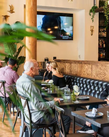 Cozy modern bistro interior with tufted black leather banquette, wooden tables, tropical plants, diners chatting over drinks and a wall-mounted TV — urban restaurant dining scene.