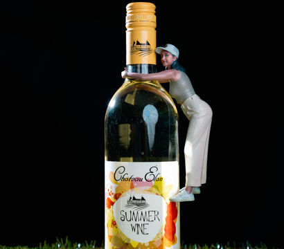 Miniature woman figurine hugging a tall bottle of summer peach white wine with a colorful floral label, set on grass against a black background.