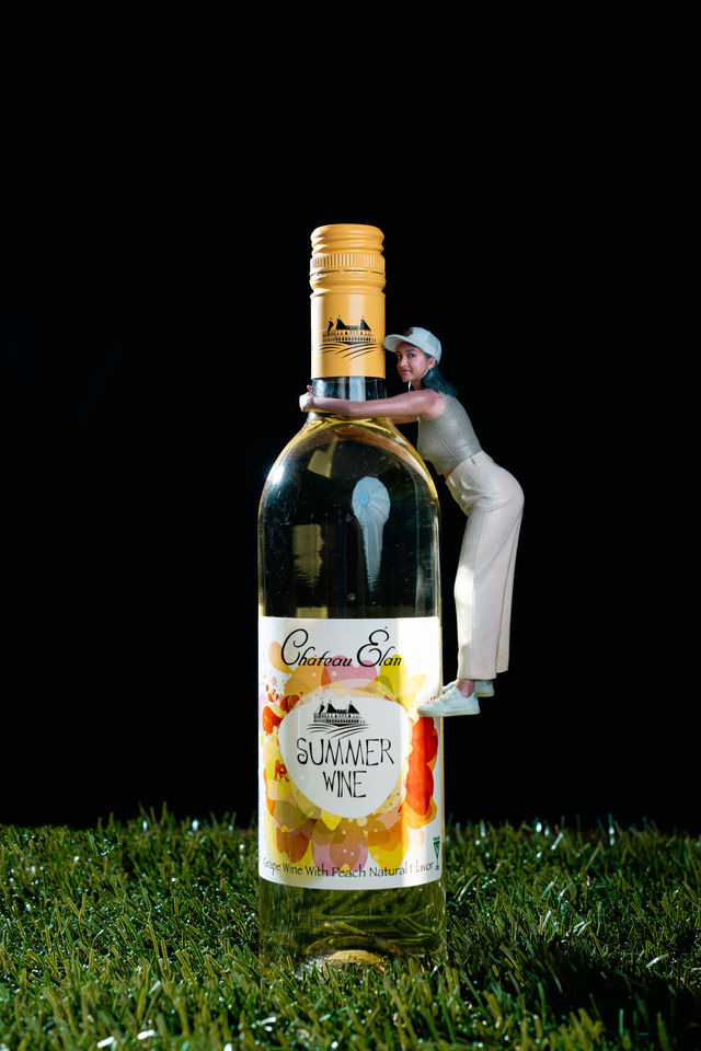 Miniature woman figurine hugging a tall bottle of summer peach white wine with a colorful floral label, set on grass against a black background.