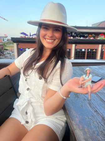 Smiling woman in a white sunhat and buttoned romper on a rooftop patio at dusk, playfully holding a tiny miniature version of herself in her hand with brick buildings and a city view behind her.