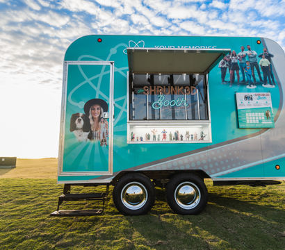 Teal mobile photo‑booth trailer with colorful people and pet graphics, open service window showing a neon 'Booth' sign and miniature figurines, parked on a sunlit grassy field under a blue, partly cloudy sky.