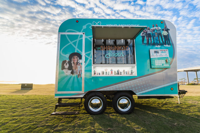 Teal mobile photo‑booth trailer with colorful people and pet graphics, open service window showing a neon 'Booth' sign and miniature figurines, parked on a sunlit grassy field under a blue, partly cloudy sky.