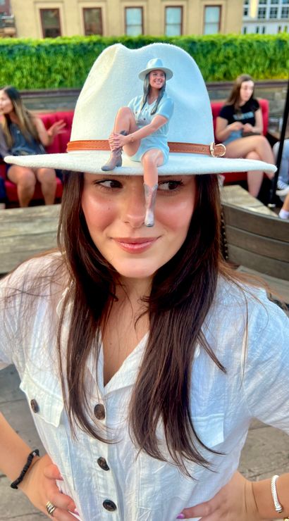 Smiling woman in a white button-up and wide-brim hat with a tiny seated figurine perched on the brim, posing on an urban rooftop bar with red-cushioned seating and city buildings in the background.
