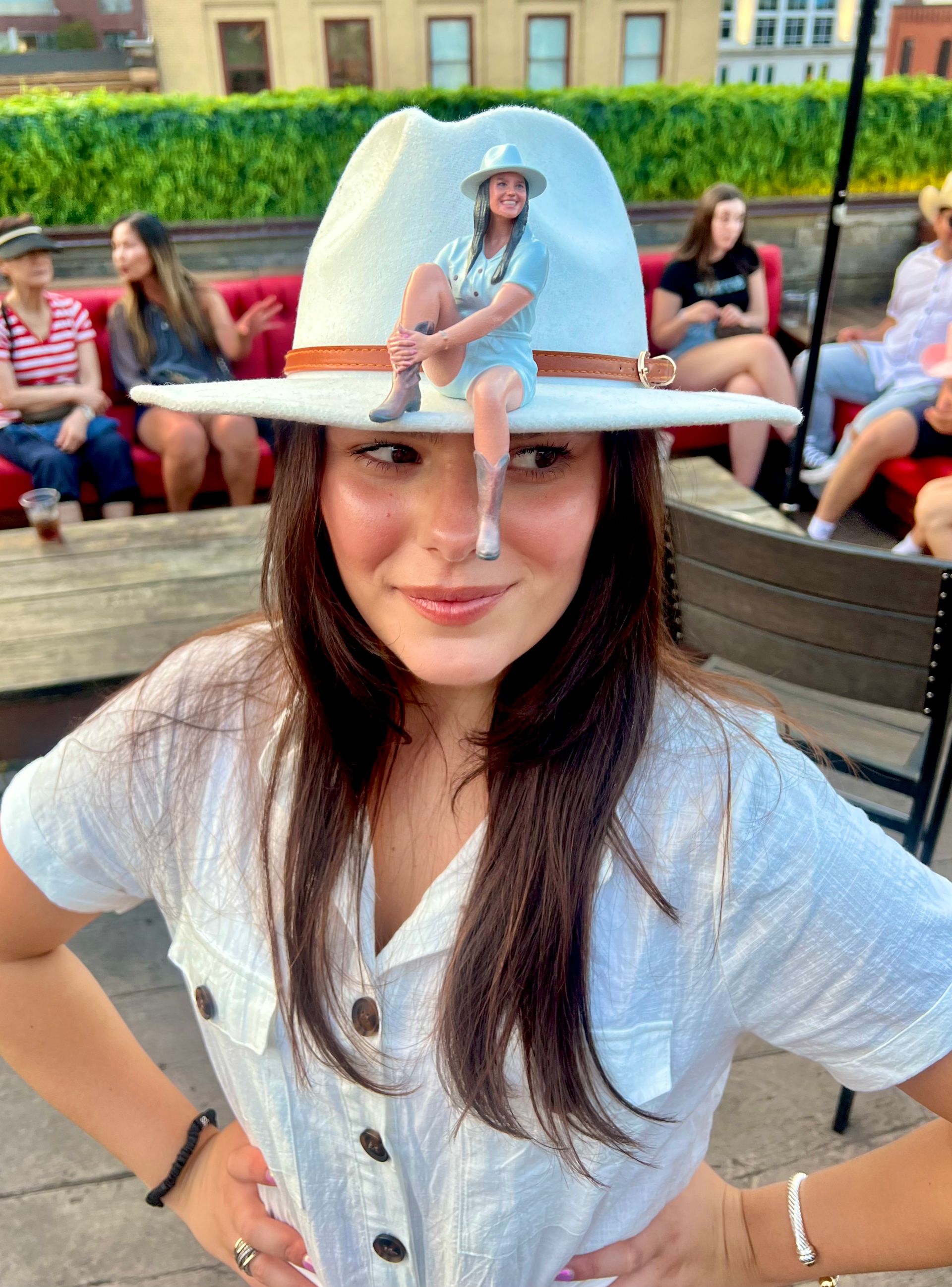 Smiling woman in a white button-up and wide-brim hat with a tiny seated figurine perched on the brim, posing on an urban rooftop bar with red-cushioned seating and city buildings in the background.