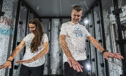 Two visitors with hands outstretched in an indoor interactive light installation, patterned projection mapping casting text-like shadows across their white t-shirts and surrounding walls.