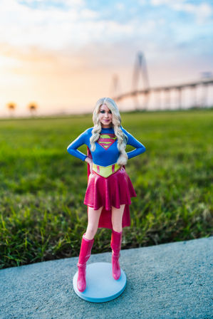 Playful superhero figurine in a blue top with S emblem, red skirt, cape and boots standing on a round base on a sidewalk by a grassy field with a suspension bridge and colorful sunset sky.