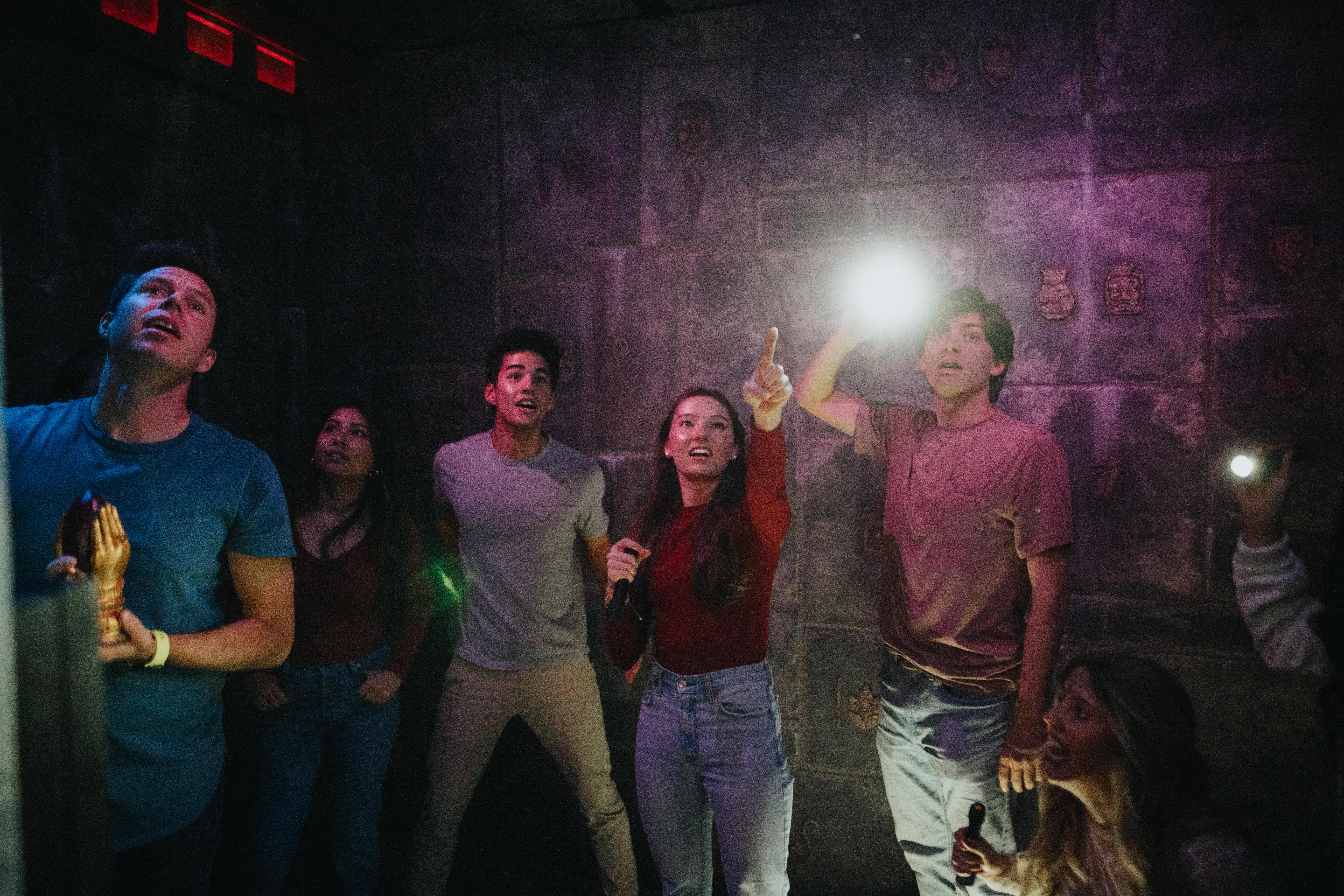 Group of six friends in a dim themed escape room inspecting carved stone walls with flashlights and excited expressions — indoor team-building puzzle game