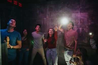 Group of six friends in a dim themed escape room inspecting carved stone walls with flashlights and excited expressions — indoor team-building puzzle game