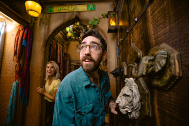 Curious man with glasses peering through an ornate wooden door with lion and elephant knockers into a narrow Moroccan-style alley, smiling woman, colorful scarves and lanterns in the background.