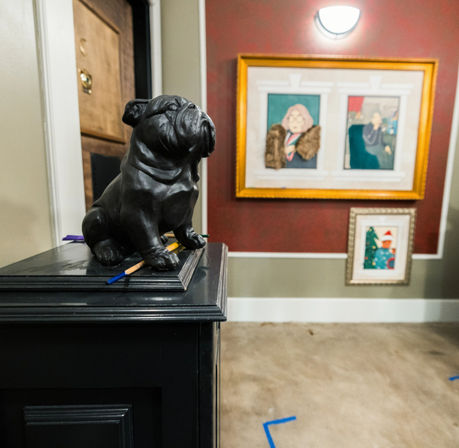Stoic black bulldog statue perched on a dark entryway cabinet with pencils, gold-framed artwork on a red accent wall and blue-tape floor markings in a home foyer.
