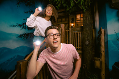 Two visitors holding flashlights and peering upward inside a dim, mountain-themed indoor treehouse exhibit with wooden railings, lantern-lit cabin backdrop and painted mountain scenery.
